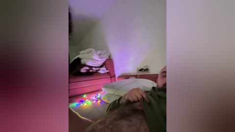 Video of a dimly lit bedroom with a person in green pajamas sitting on the floor, holding a white plate, beside a bed with a white comforter, and a string of colorful lights.