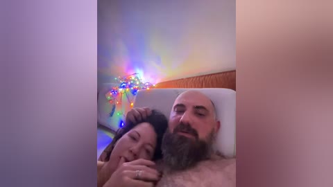 A video showing a bald man and a woman with long hair, both shirtless, lying in bed under a rainbow-colored string of lights.
