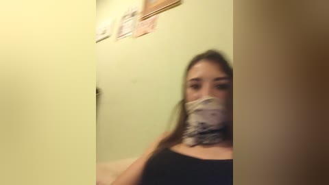 A blurry video of a young woman with light skin, brown hair, wearing a black top and a patterned face mask, taken in a dimly lit room with yellow walls.
