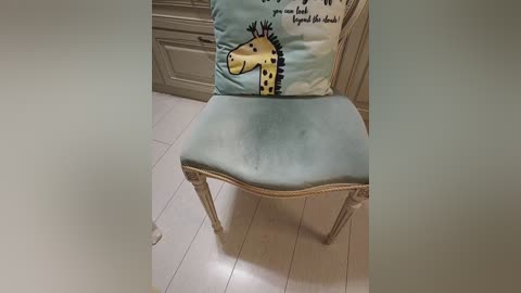 Video of a vintage, light blue upholstered chair with a cushion featuring a giraffe illustration on a light blue background, positioned on a light wooden floor, with a white door in the background.