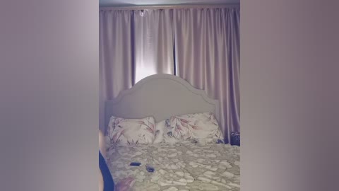 Video of a bedroom with a large, ornate headboard and a bed covered in a floral-patterned duvet, surrounded by light pink curtains.