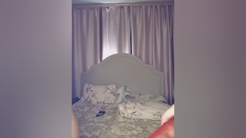 A video of a bedroom with a white, upholstered headboard and light-colored bedding featuring a floral pattern. The background features a large window with beige curtains partially drawn. The image appears slightly out of focus.