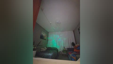 Video of a dimly lit, cluttered bedroom with a single bed covered in a black blanket, a colorful backpack on the floor, and a white curtain with green light streaming through.
