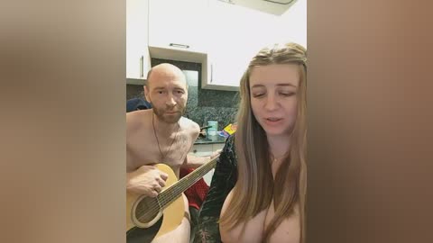 Video of a bald, shirtless man playing an acoustic guitar, accompanied by a topless young woman with long hair, in a modern kitchen with white cabinets and black countertops.