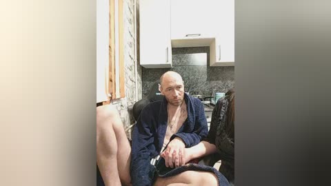 Video of a bald, middle-aged man in a robe, sitting on a kitchen counter, legs apart, in a modern kitchen with white cabinets and a grey marble backsplash.