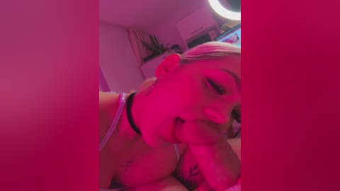 Video of a young woman with light skin and platinum blonde hair, wearing a black choker, lying on a bed, partially obscured by a pink filter, in a dimly lit room with a round light.