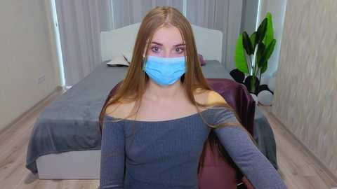 Video of a young Caucasian woman with light skin, long straight blonde hair, wearing a blue face mask, off-shoulder gray sweater, and seated on a red chair in a modern, minimalistic bedroom with gray bedding, white walls, and a green plant.