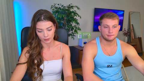 Video of a young Caucasian couple: a brunette woman in a white tank top, and a blond man in a blue tank top. They sit on a black office chair in a modern office with a green plant, blue lighting, and a TV on the wall.