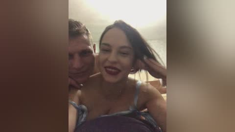 Video of a smiling woman with dark hair, wearing a low-cut blue top, leaning against a man with short hair in a dimly lit room.