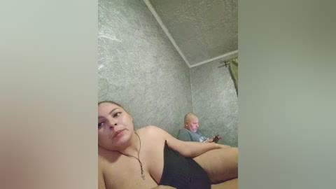 Video of a young, topless woman with medium breasts, lying on a bed, while an older man with glasses, in a gray shirt, sits nearby.