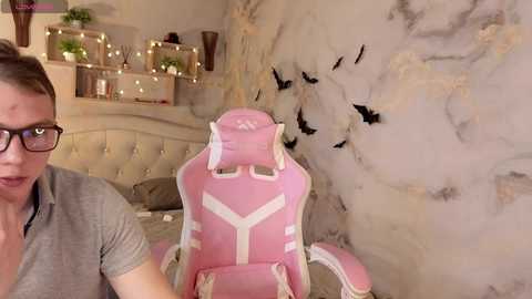 Video of a young woman with glasses, light skin, and short brown hair, sitting in a pink gaming chair with a white geometric design. Behind her, a marble-patterned wall with black bats is visible.