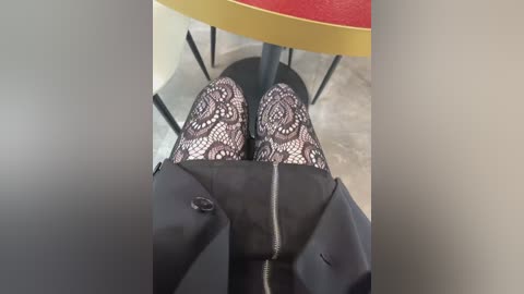 Video of a person sitting at a table with black lace-patterned stockings, black jacket, and silver zipper details. The background features a red and yellow round table, black chairs, and a light-colored floor.