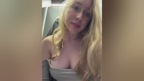 Video of a young Caucasian woman with long blonde hair, fair skin, and a light grey off-shoulder top, revealing ample cleavage. She is in a dimly lit bathroom with a beige towel hanging on a rack in the background.