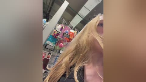 Video of a blonde woman with shoulder-length hair, partially cropped from the top, standing in a cluttered, dimly-lit store aisle. Background shows colorful, disorganized shelves with products.