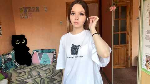 Video of a young white woman with long brown hair, wearing a white T-shirt with a black dog graphic and text, standing in a cozy, colorful room with a black cat and patterned couch.