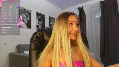 Video of a smiling woman with long blonde hair and fair skin, wearing a pink strapless top, sitting in a black leather chair in a modern, minimalist room with black-and-white floral artwork and a digital TV screen displaying a menu.