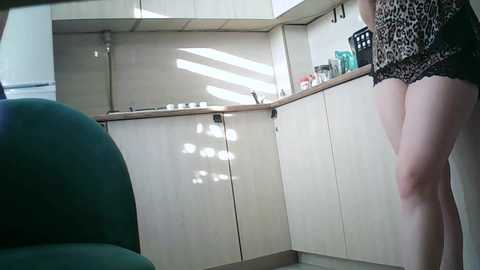 Video of a woman in leopard print shorts standing in a modern kitchen with light wood cabinets and white counters. A green chair is partially visible on the left.