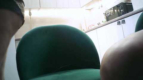 A video of a kitchen with a green velvet chair in the foreground, white cabinets and appliances in the background, featuring a microwave and a countertop filled with various kitchen items.