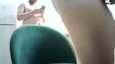 Video of a topless man with a beard and light skin, wearing a red necklace, standing in a kitchen. The image is slightly blurry, showing a green chair and a refrigerator in the background.