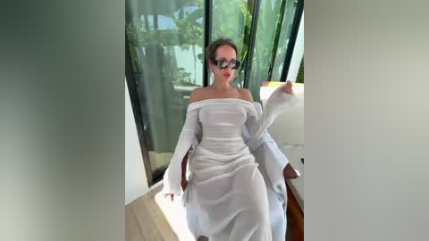 A video shows a woman in a white off-shoulder dress, with glasses, sitting on a white chair in a modern room with large windows and greenery outside.