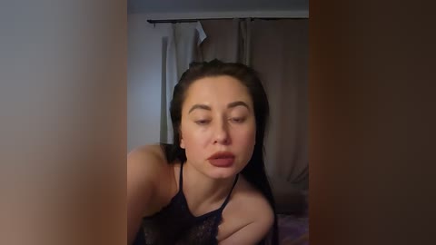 A video of a young woman with fair skin and dark hair, wearing a black lace top, looking contemplative in a dimly-lit bedroom with beige curtains.