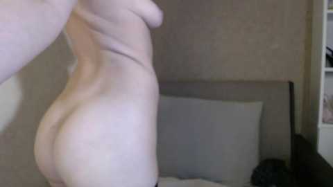 A video of a nude, light-skinned person with a slender, toned physique standing sideways in a dimly lit bedroom. The background features a beige wall and a partially visible bed with white sheets.