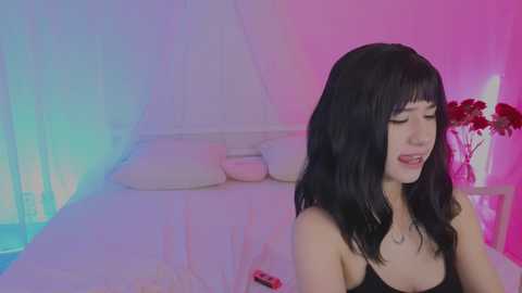 A video of an Asian woman with long black hair, wearing a black tank top, in a softly lit room with pink and blue hues.