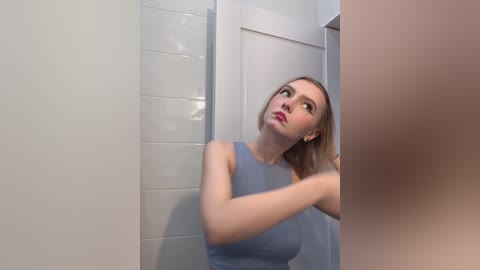 Video of a blonde woman in a light blue dress, holding a phone in a white-tiled bathroom.