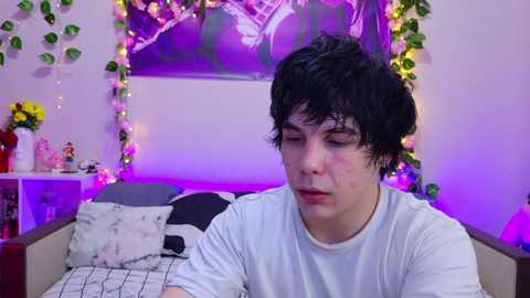 Video of a young man with curly black hair, wearing a white T-shirt, sitting on a bed with a floral-patterned pillow. Background features purple string lights, a framed art piece, and a white shelf with decorations.