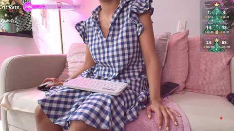 Video of a light-skinned woman with a slender build wearing a blue and white gingham dress, sitting on a pink and white striped couch, typing on a white keyboard, in a cozy, softly lit room.