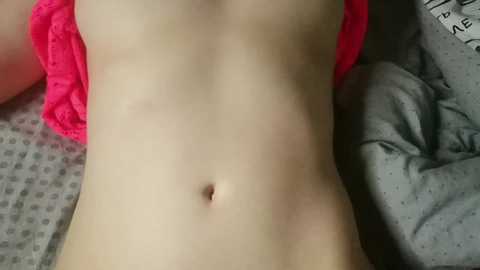 Video of a pale, slim, nude torso with small breasts, lying on a gray and white polka-dotted bedspread. A bright pink garment is partially visible on the right side.