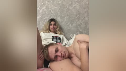 A video of two young women in a dimly lit room. One, blonde, sits with a hoodie on, while the other, brunette, lies naked on her back. The background has a textured, light-colored wall.