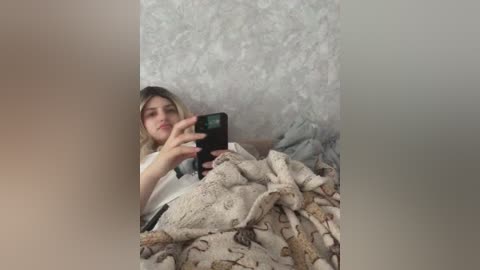 Video of a young woman with light skin and blonde hair, lying in bed with a beige patterned blanket. She holds a smartphone, taking a selfie against a textured wallpaper background.