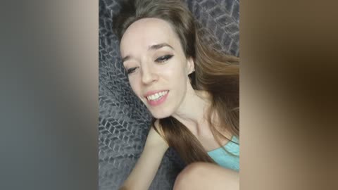 Video of a smiling, fair-skinned woman with straight brown hair, wearing a turquoise tank top, lying on a textured gray blanket. The image is cropped, with a blurred, brown-toned background.