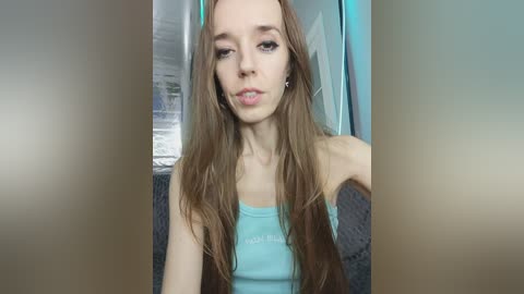 Video of a young, slender, Caucasian woman with long, straight brown hair, wearing a light blue tank top. She has a neutral expression and is indoors, possibly in a living room with a grey couch and a white window in the background.