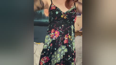 A video of a woman in a black floral dress with spaghetti straps, standing in a dimly lit room with a blurred background of a bed and a wooden dresser.