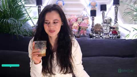 Video of a young woman with long black hair, fair skin, and a white top, holding a glass of whiskey, in a modern living room with plants, plush toys, and a black sofa.