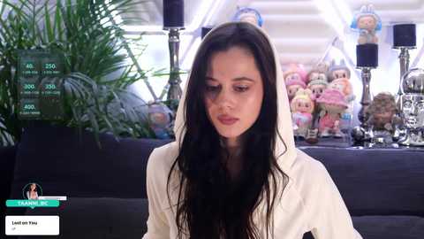 Video of a young woman with long, dark hair, wearing a white robe, sitting in a modern living room with a black couch, plush toys, and greenery.