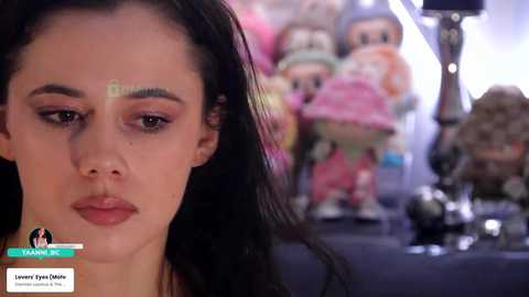 Video of a young woman with dark hair, fair skin, and minimal makeup, looking thoughtful, in front of a colorful toy display featuring various cartoon characters.