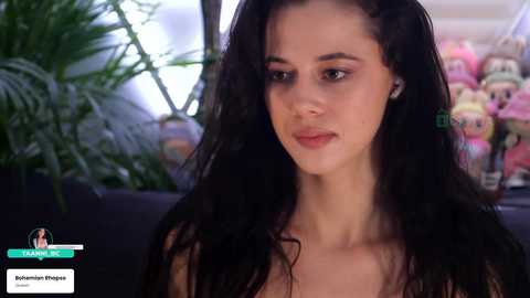 A video of a young woman with long, dark hair and fair skin, wearing minimal makeup, sitting indoors with a plush toy background.