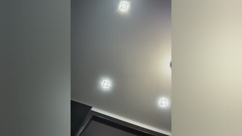 Video of a modern ceiling with four recessed, square-shaped lights emitting a soft glow. The ceiling is painted in a light gray color, contrasting with the dark trim below. The image is well-lit, highlighting the minimalist and sleek design.