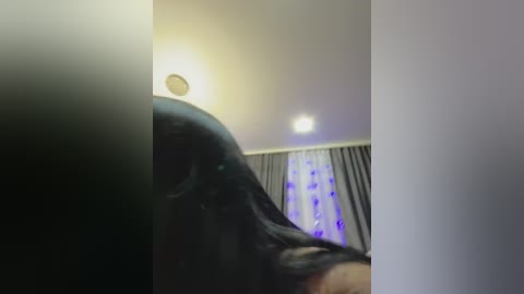 A blurred video shows a person with long, dark hair partially visible. The background features a white wall with a light fixture and a curtain with a purple pattern.
