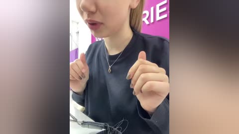 Video of a fair-skinned woman with light brown hair, wearing a black lab coat, making thumbs-up gestures, in a bright, modern room with purple and white walls.