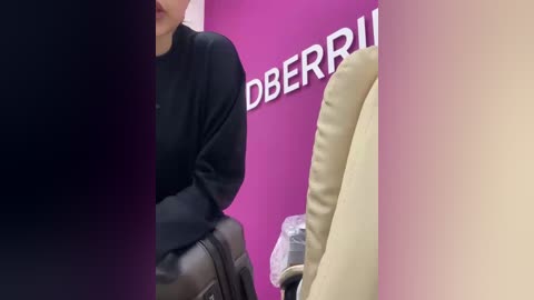 Video of a person with fair skin wearing a black top, sitting on a beige leather chair against a pink wall with \"BBRUM\" in white letters.