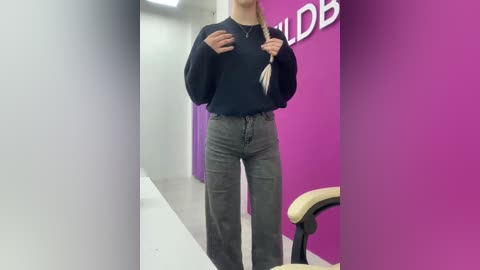 A video of a woman standing in a brightly lit room with purple walls. She wears a black sweater and gray high-waisted pants, holding a feather.