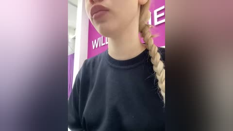 Video of a young woman with light skin and long blonde hair in a braid, wearing a black sweatshirt, standing in front of a partially blurred purple wall with white text.