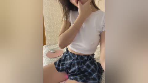 Video of a young woman with long dark hair, wearing a white cropped top and blue plaid mini skirt, sitting on a bed with beige wallpaper in the background.