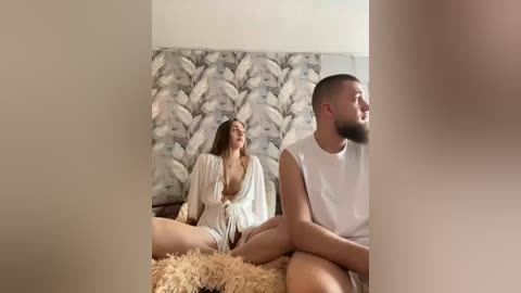 Video: A light-skinned, bearded man in a white tank top sits on a bed, gazing at a fair-skinned woman in a revealing white robe, sitting on a fluffy rug in a minimalist bedroom with leaf-patterned wallpaper.