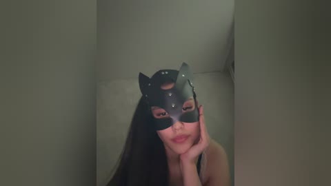 Video of a woman with long black hair, wearing a black cat mask, leaning on her hand, in a dimly lit room.