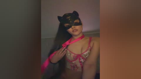 Video of a woman in a black cat mask, pink lingerie, and a pink ribbon, standing indoors with a blurred background.